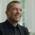 Ein Mann in einem schwarzen Anzug lächelt, mit einem unscharfen, hellen Fenster im Hintergrund.