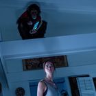 Ein Affe mit Tablet sitzt oben, während eine junge Frau unten steht, beide scheinen überrascht in einem Raum.