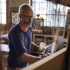 Ein Maler mit Brille im Atelier, arbeitet an einer Leinwand, hielt ein Glas mit Pinsel in der Hand, umgeben von Porträts.
