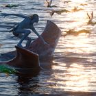 Illustration eines blauen Wesens auf einer Kreatur im Wasser, umgeben von fliegenden, vogelartigen Geschöpfen bei Sonnenuntergang.