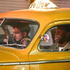 Text: "Yellow" und "VACANT." Zwei Männer sitzen in einem alten gelben Taxi; einer fährt, der andere ist Beifahrer.