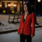 Eine Frau steht nachts draußen, gekleidet in einen leuchtend roten Blazer. Im Hintergrund scheint Licht aus einem Haus.