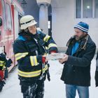 Zu sehen ist die Zahl 112 auf einem roten Feuerwehrauto. Feuerwehrleute übergeben etwas an zwei Männer im Schnee.