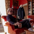 Ein älterer Mann sitzt in einem roten Sessel mit einem Glas. Ein weiterer Mann steht im Hintergrund mit einem dunklen Mantel.