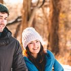 Ein Mann und eine Frau lächeln im Winter, beide tragen Mützen und Jacken, Bäume im Hintergrund.