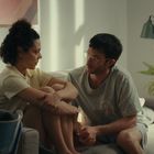 Zwei Personen sitzen auf einem Sofa, schauen sich an. Im Hintergrund ist ein abstraktes Kunstwerk und eine Pflanze.