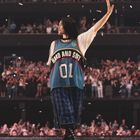 Eine Person in einem Jersey mit "HARD AND SOFT 01" hebt die Hand in einer Arena voller jubelnder Menschen.
