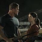 Ein muskulöser Mann und eine Frau stehen nah beieinander, ernst schauend, er hält ein Gewehr. Im Hintergrund verschwommen.