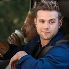 Ein junger Mann sitzt in einem Auto, lächelt und schaut aus dem Fenster. Ein älterer Mann trinkt im Hintergrund Kaffee.