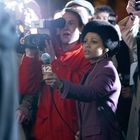 „12“ auf einem Mikrofon. Eine Reporterin mit Mikrofon und Kamera umgeben von Journalisten. Nachtaufnahme.