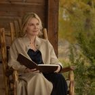 Keine Texte vorhanden. Eine Frau in einem hellen Mantel sitzt draußen auf einem Holzstuhl und liest ein Buch.