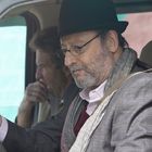 Ein Mann mit Brille und Hut sitzt am Steuer eines Autos. Ein anderer Mann sitzt daneben.
