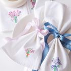 Bestickte Servietten mit floralen Motiven, rosa und blauen Schleifen, Tasse und Untertasse im Hintergrund.