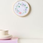 Ein Stickrahmen an der Wand mit einem Blumenkranzmotiv. Darunter ein Buchstapel und eine beige, runde Vase.