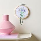Gesticktes Blumenmotiv in einem runden Stickrahmen, daneben eine pinke Vase auf einem hellen Regal.