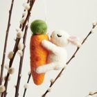 Ein Filzhäschen umarmt eine große Karotte. Im Hintergrund sind Zweige mit flauschigen Knospen.