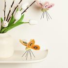 Zwei Wollschmetterlinge, ein orangefarbener und ein rosafarbener, umgeben von weißen Tulpen in einer Vase auf einem Regal.