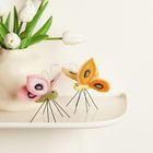 Zwei filzartige Schmetterlinge in Rosa und Orange stehen neben einer Vase mit weißen Tulpen.