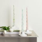 Drei dekorative, lange Kerzen mit floralen Mustern auf einem Metalltisch; daneben weiße Tulpen.