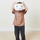 Ein Kind hält einen weißen Korb mit Hasengesicht und Ohren vor dem Gesicht, trägt ein braunes Shirt und dunkle Jeans.