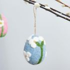 Ein blauer Filz-Osterei-Anhänger mit weißen Blumen hängt an einem Weidenzweig. Links im Hintergrund ist ein pinker Anhänger.