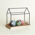 Keine Texte vorhanden. Ein Drahtgestell in Hausform mit Filzeiern und Stroh darin auf einer Holzbasis.