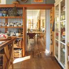 AUGUST 2027. Ein gemütliches Haus mit Holzböden, einem Essbereich und Regalen voller Geschirr und Töpfen.