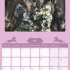 „Mai“ steht auf einem lilafarbenen Kalender. Zwei schwarze Pferde stehen vor blühenden Bäumen.