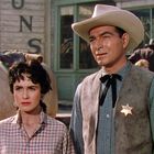 Ein Sheriff in Westernkleidung mit einem Sternabzeichen steht neben einer Frau in einem karierten Hemd.