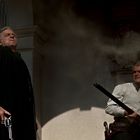 Zwei Männer mit Waffen stehen vor einem Gebäude. Der linke Mann in schwarz trägt einen Revolver, der rechte ein Gewehr.