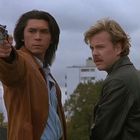 Zwei Männer im Freien; einer mit erhobenem Revolver. Der Himmel ist bewölkt, Laubbäume im Hintergrund.