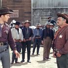 In einer Westernszene steht links ein Mann mit Sheriff-Abzeichen vor einer Gruppe von Männern; sie tragen Cowboyhüte.
