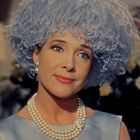 Eine Frau mit blauer Perücke und Perlenkette lächelt leicht. Im Hintergrund sind Blumen zu sehen.