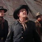 Drei Männer in Cowboy-Kleidung stehen aufmerksam in einer felsigen Wüstenlandschaft, im Hintergrund sind Pferde zu sehen.