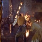 Ein brennender Stall mit zwei Menschen, die eine Kuh retten. Flammen lodern, Holz und Wagenrad im Hintergrund.