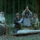 Vier Personen in mittelalterlicher Kleidung; eine hält ein Schwert über einer eingewickelten Gestalt liegend. Wald im Hintergrund.