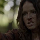 Eine Frau mit langen Haaren und entschlossenem Blick zielt im Wald mit einer Pistole. Ihr Gesicht zeigt Schmutz und Verletzungen.