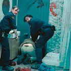 Zwei Polizisten in blauen Uniformen beugen sich über eine am Boden liegende dunkel gekleidete Person in einem blutverschmierten Raum.