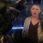 Eine Frau hinter Glas, umgeben von zwei Dinosaurierfiguren. Sie schaut überrascht. Im Hintergrund Pflanzen.