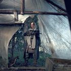 Person in einem improvisierten Unterstand aus Holz und Plastikplanen. Sie trägt warme Kleidung und hält eine Waffe.