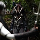 Eine dunkel gekleidete Gestalt mit Knochen-Accessoires steht im Wald. Im Vordergrund eine Person mit weißer Bemalung.