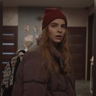 Eine Frau mit roter Mütze und gepolsterter Jacke steht in einem Flur, trägt einen gemusterten Rucksack.