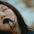 Das Gesicht einer Person mit blassen Augen, dunklen Lippen und pechschwarzen Linien auf blasser Haut im Freien.