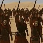 Personen tragen Masken und braune Roben, halten Stäbe, in einer Wüste mit Hügeln im Hintergrund.