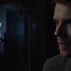 Ein junger Mann schaut ernst, im Hintergrund steht eine schattenhafte Figur auf einer Treppe.