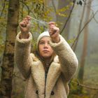 Ein Mädchen mit weißer Mütze und flauschigem Mantel im nebligen Wald hält ein Spinnennetz in der Hand.