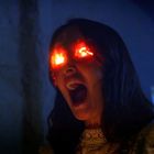 Eine Frau mit entsetztem Gesichtsausdruck, aus deren Augen ein helles, rotes Licht strahlt.