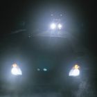 Autoscheinwerfer leuchten in der Dunkelheit direkt auf den Betrachter, ein weiteres Fahrzeug leuchtet im Hintergrund.