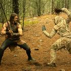 Zwei Figuren in einem Wald: links ein Krieger mit Schwert, rechts ein mumienartiges Wesen mit einer Keule aus Metall.