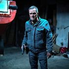Text "DISCO" im Hintergrund. Ein älterer Mann in blauer Jacke hält eine Waffe, im Dunkeln neben einem roten Boot.